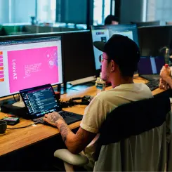 Jovem sentado mexendo no computador. Ele usa uma camisa branca, óculos e um boné preto. Sua pele é branca e seus cabelos são pretos.