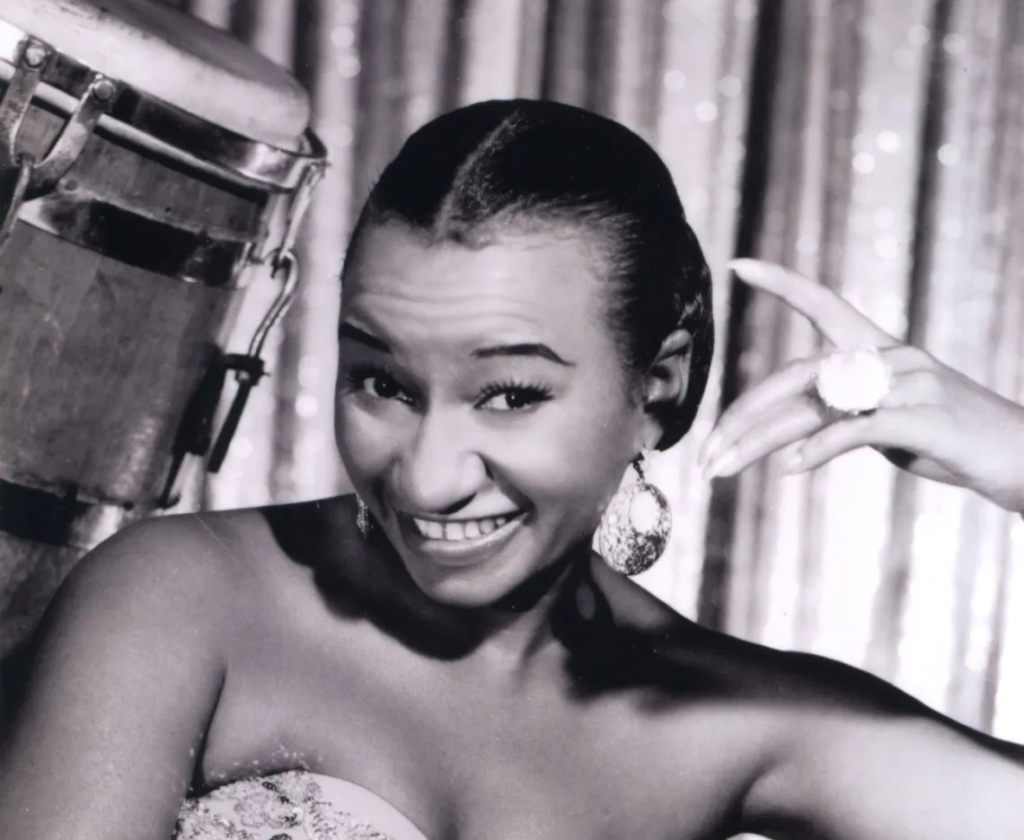 Celia Cruz posa para foto en blanco y negro, joven con pendientes y un gran anillo en el dedo de la mano izquierda, sonríe.