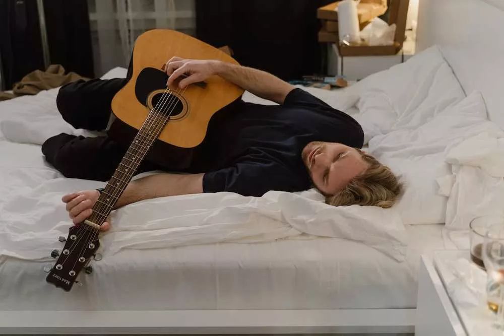 Hombre acostado en una cama con sábanas blancas tiene una guitarra acústica en las manos y parece sin motivación para tocarla