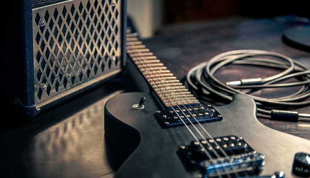 Guitarra preta apoiada no chão, ao lado de um cabo e um amplificador