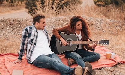 Mulher tocando violão para o namorado, com os dois sentados em uma canga estendida na natureza a céu aberto