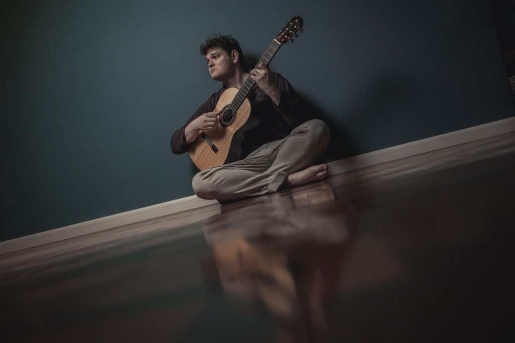 Lucas Telles com violão em mãos e sentando no chão de uma sala