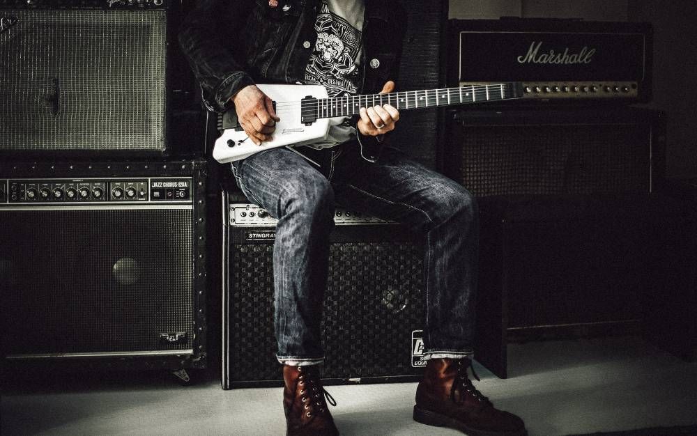 Guitarrista sentado à frente de uma parede de modelos de amplificador de guitarra