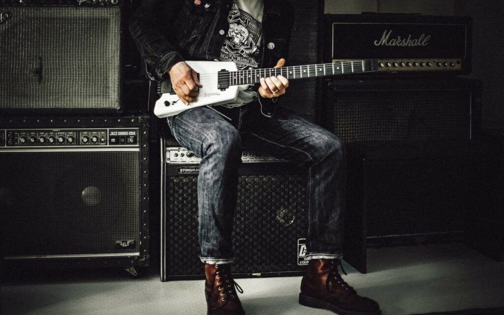 Guitarrista sentado em frente a uma parede de amplificadores de guitarra
