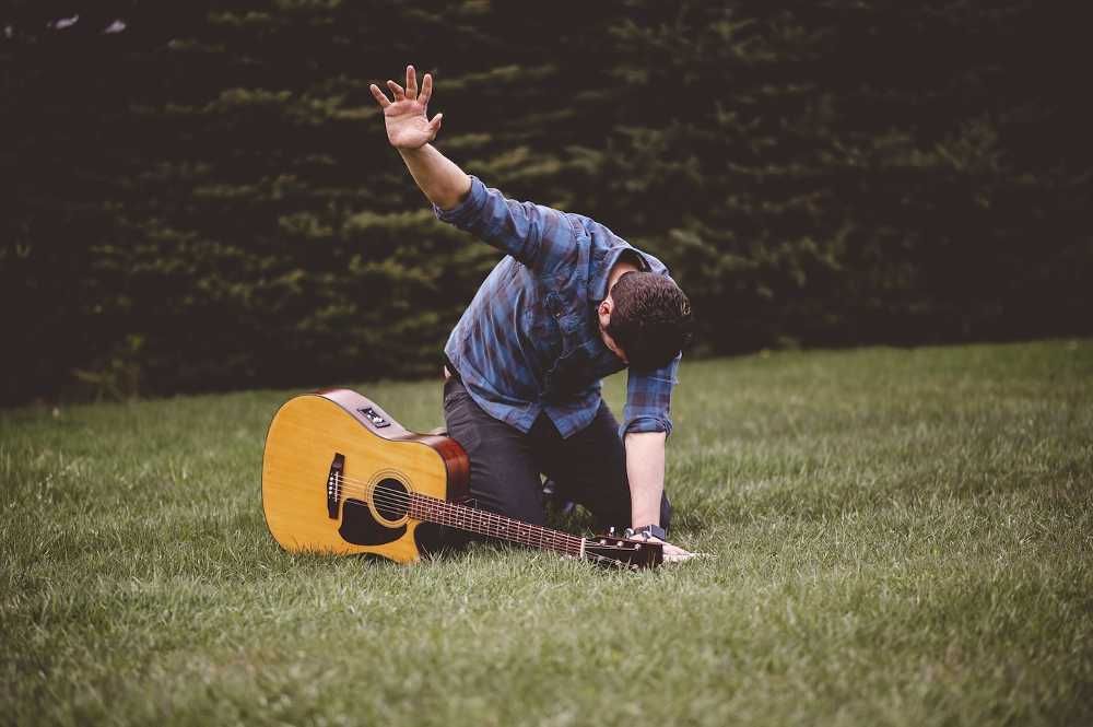 Homem ajoelhado na grama com um violão e estendendo mão direita ao céu