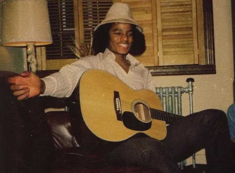 Michael Jackson jovem com um violão no colo, com o braço direito esticado, a mão aberta e vestindo um chapéu