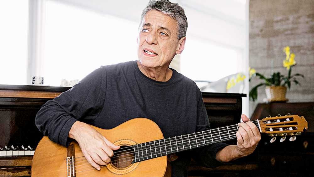 Chico Buarque tocando violão em casa e cantando olhando para o lado