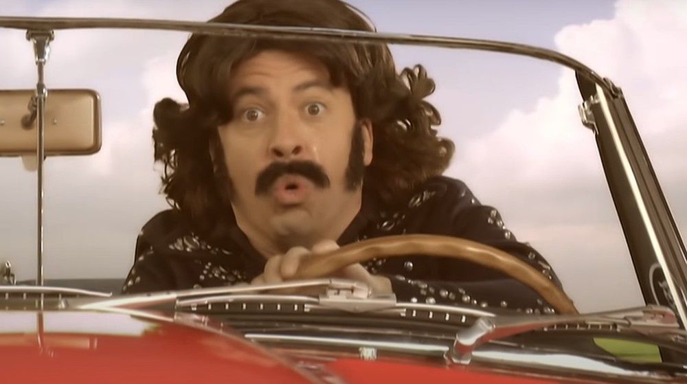 Dave Grohl no clipe de Long Road To Ruin caracterizado com bigode e peruca dirigindo carro