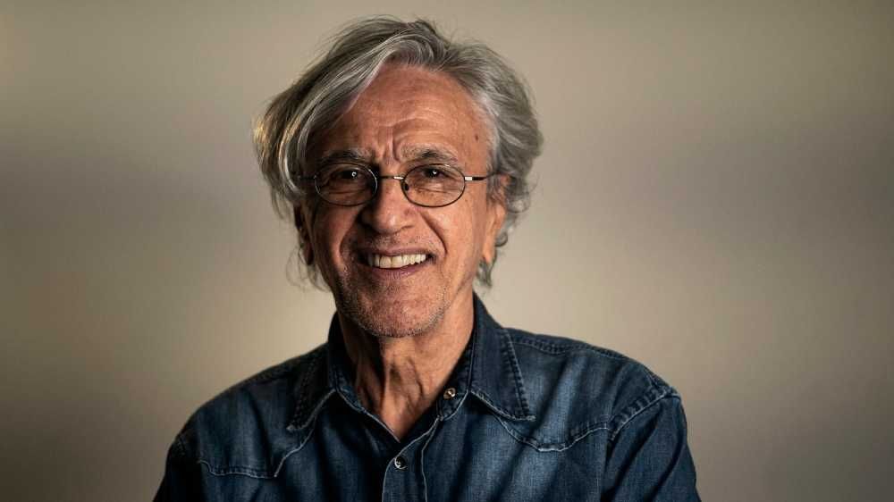 Caetano Veloso em sessão de fotos olhando para a câmera sorrindo e de jaqueta jeans