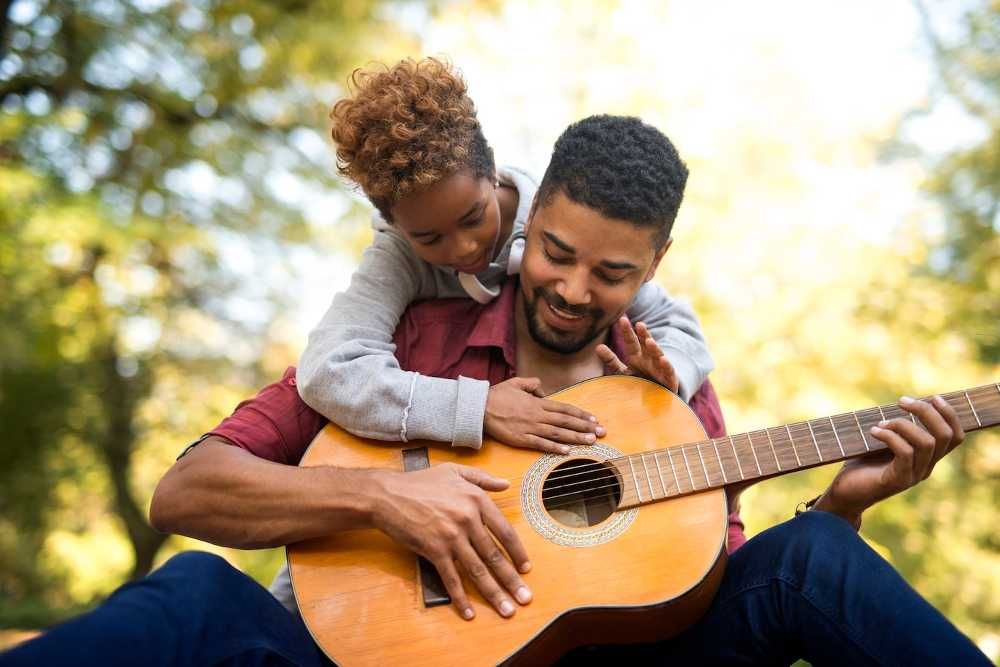 Homem tocando violão e a filha pequena segurada em suas costas com as mãos no instrumento