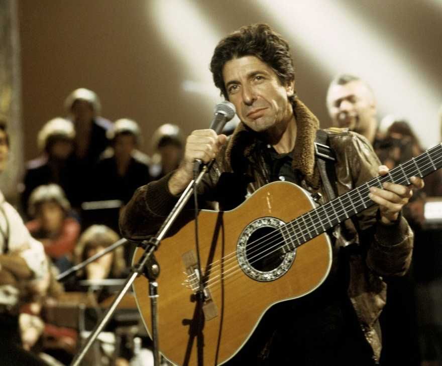 Leonard Cohen com violão no colo e segurando microfone em show