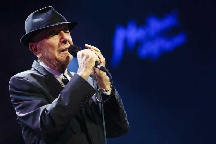 Leonard Cohen cantando de chapéu, de olhos fechados e segurando o microfone com as duas mãos em show