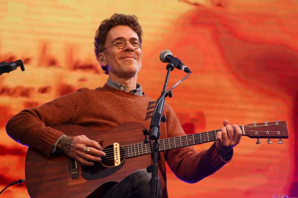 Nando Reis tocando violão em show sentado em frente ao microfone e olhando feliz para o público