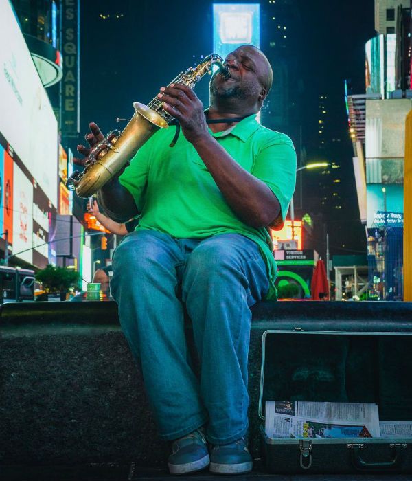 Homem negro, gordo e mais velho toca saxofone, sentando em um banco de praça
