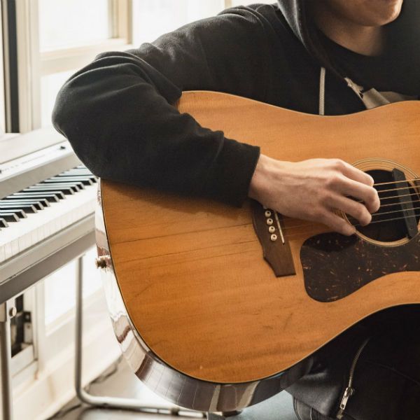 Jovem sentado no banco de um home studio toca violão. Atrás dele, há um teclado