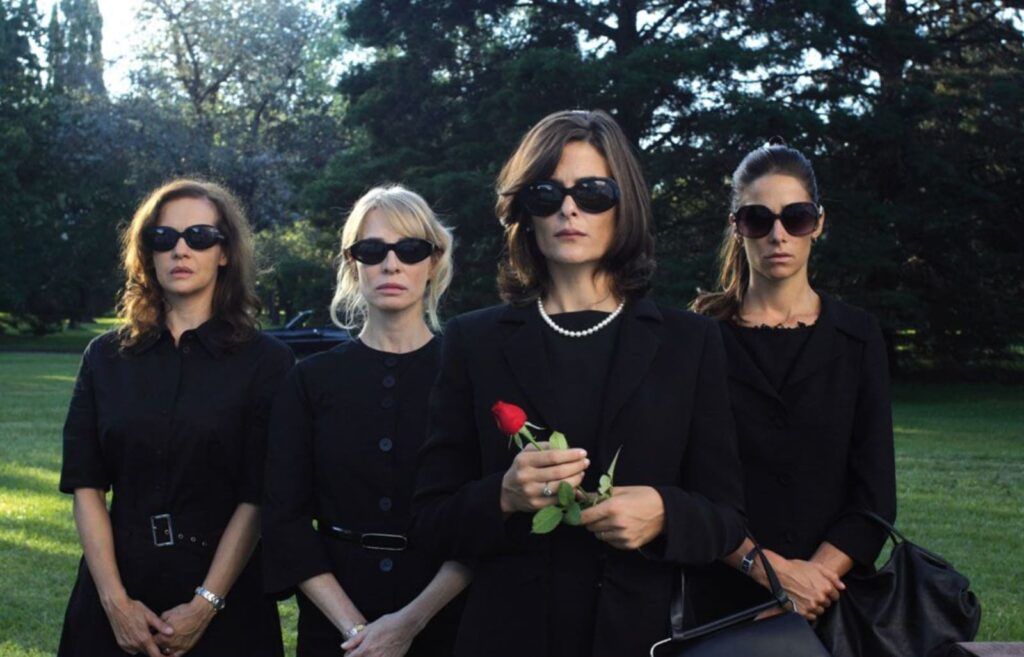 Cuatro mujeres de traje negro y gafas oscuras en un cementerio