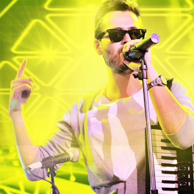 Cantor Edward Maya canta com a mão direita apontada para cima no centro da imagem