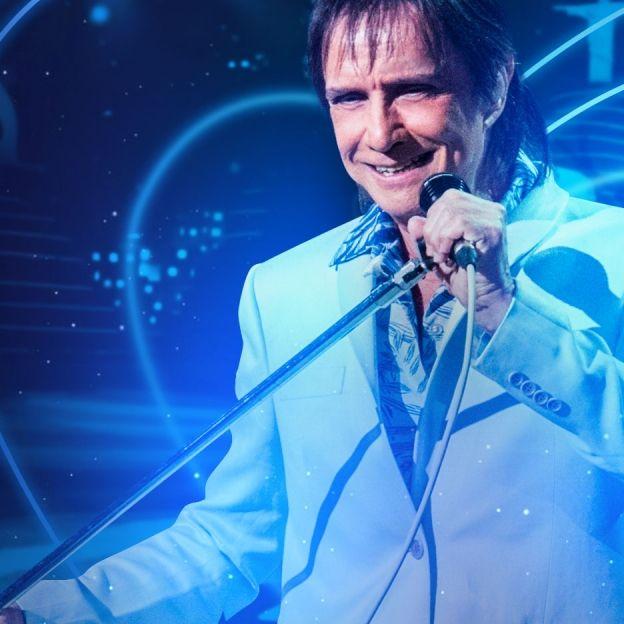 O cantor Roberto Carlos canta com um microfone na mão esquerda no centro da imagem