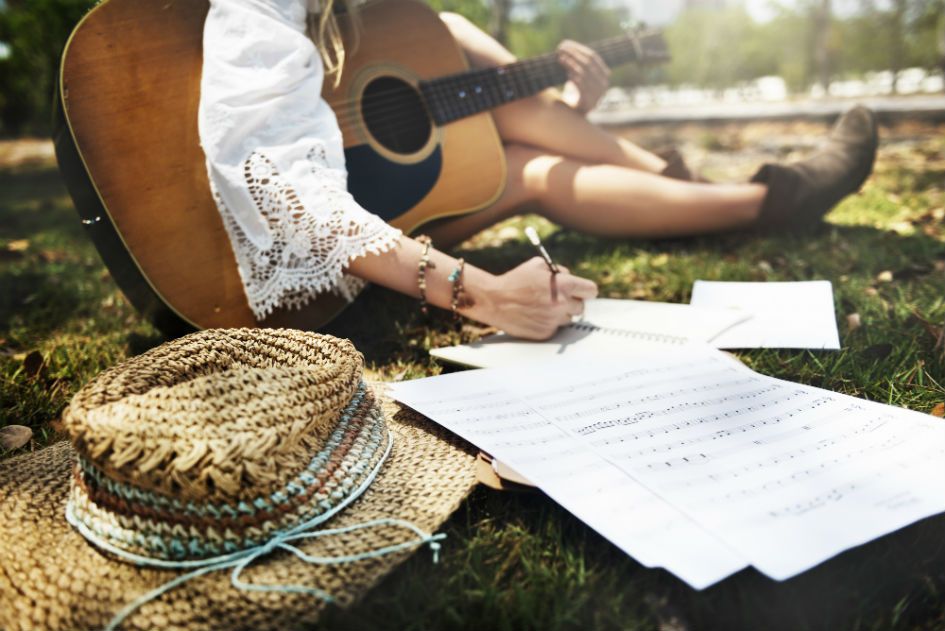 Com violao no colo, mulher escreve musica sentada na grama de um jardim