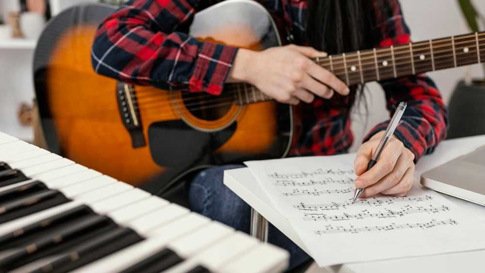 Estudante de música em frente a um teclado com um violão no colo e anotando em uma partitura